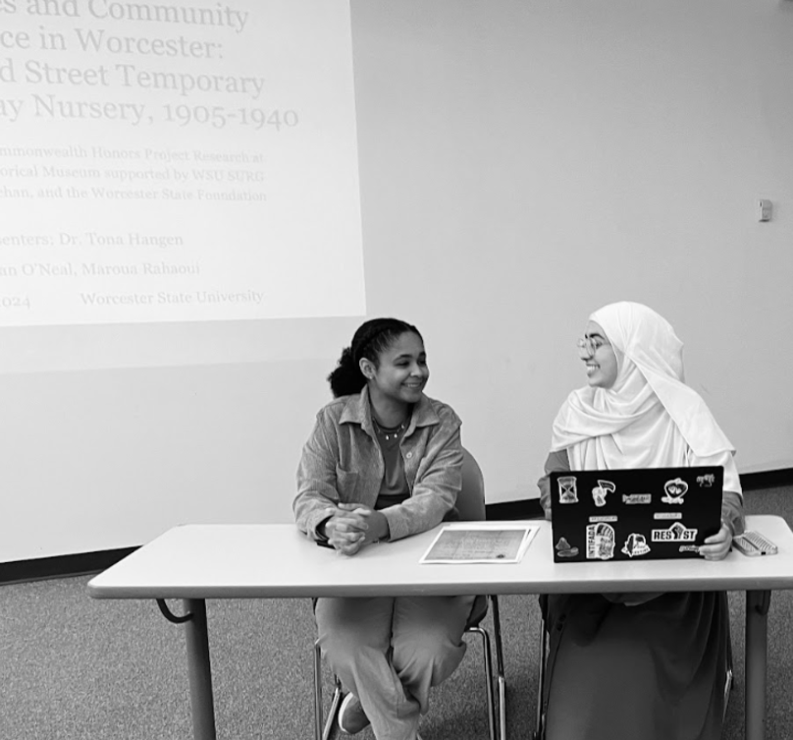 black and white photo of two woman, (Tiernan O’Neal and Maroua Rahaoui), sitting at a table with a laptop looking at each other smiling