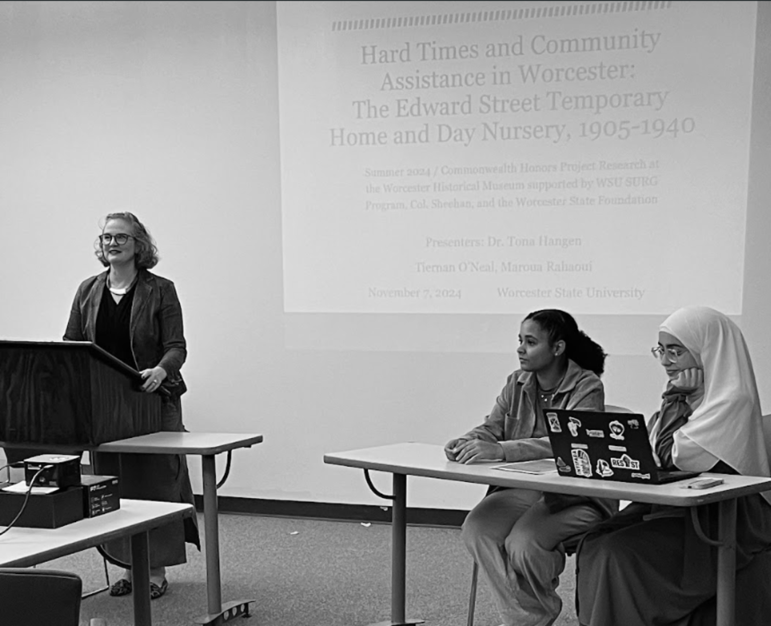 Black and white photo of a woman (Tona Hangen) standing at a podium on the left, and two woman ((Tiernan O’Neal and Maroua Rahaoui) sitting at a table with a laptop to the right. A presentation projected on the wall behind them.