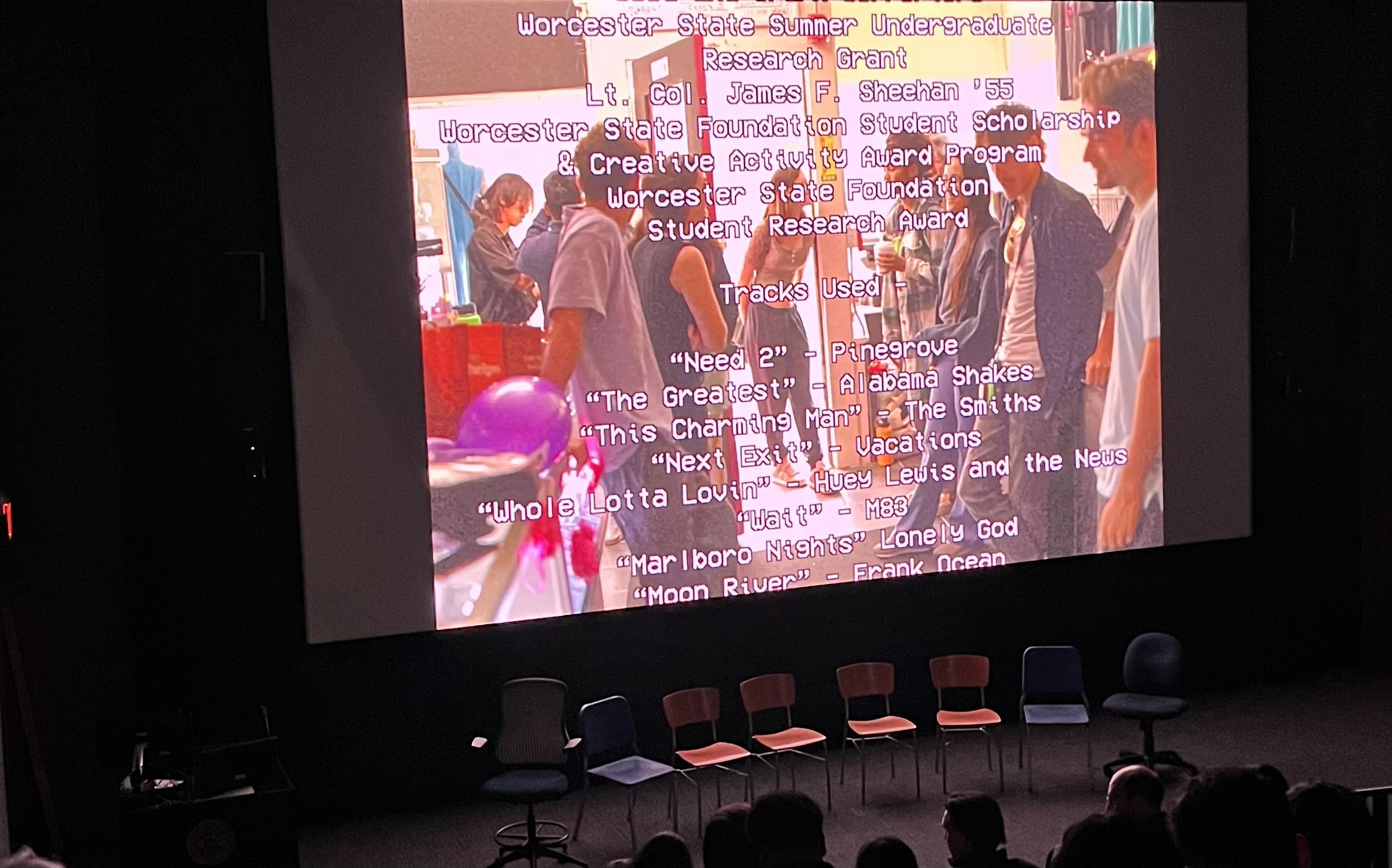 Film screening in dark cinema setting with credits rolling and empty chairs in front of screen facing audience