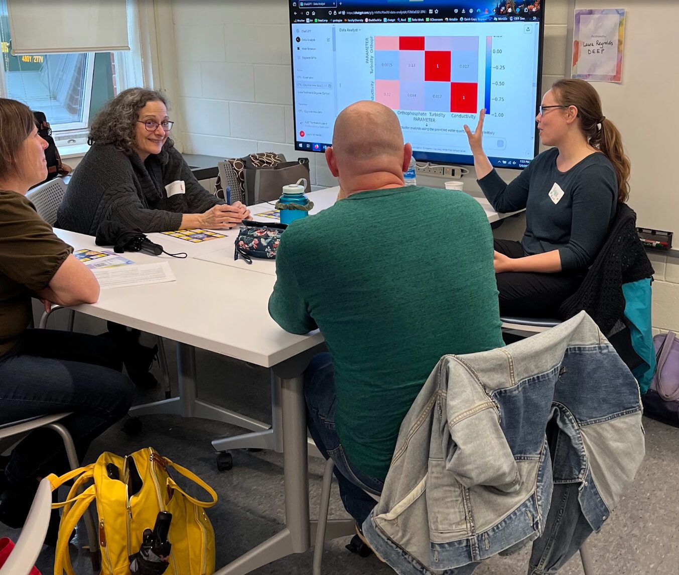 3 faculty of different ages, race, and gender seated at a table in front of a big screen with data graphs listening to a white woman with long brown ponytail and glasses teach them.