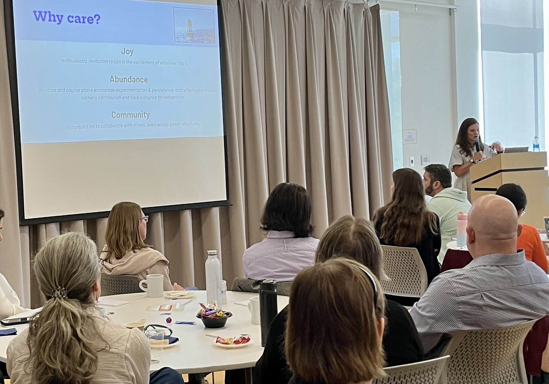 screen displaying the words joy, abundance, community and the backs of 8 faculty heads listening attentively to a white middle age woman with dark long hair speaking at podium