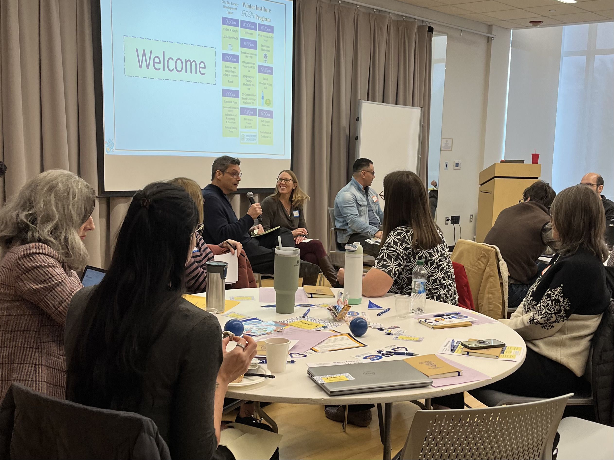 A group of faculty members sitting at a round table during a workshop, listening and gesturing as one colleague speaks.
