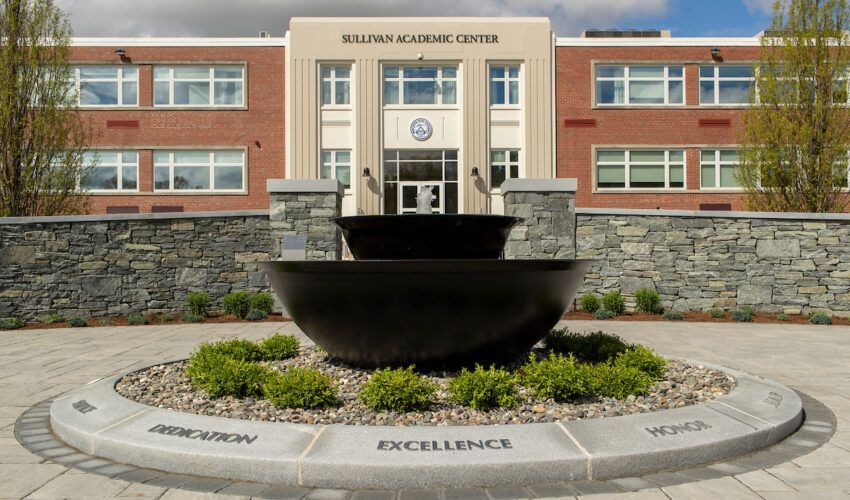 A large black fountain stands in front of the Sullivan Academic Center, home to the Center for Teaching & Learning: Faculty Development Center, with “Dedication,” “Excellence,” and “Honor” engraved on the surrounding walkway.