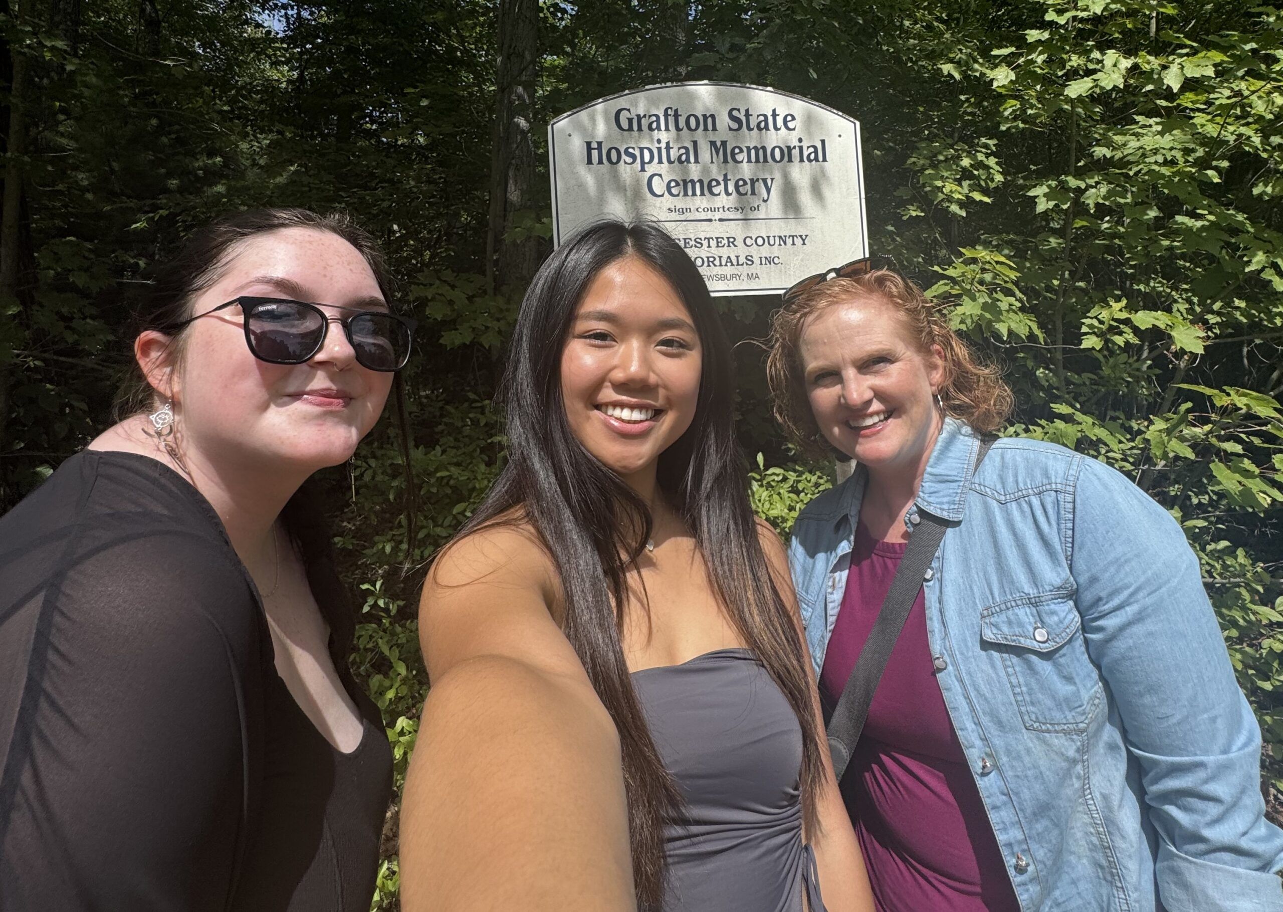 Three women, participants in the Summer Undergrad Research Grant Program, stand smiling before a sign reading "Grafton State Hospital Memorial Cemetery," surrounded by trees.