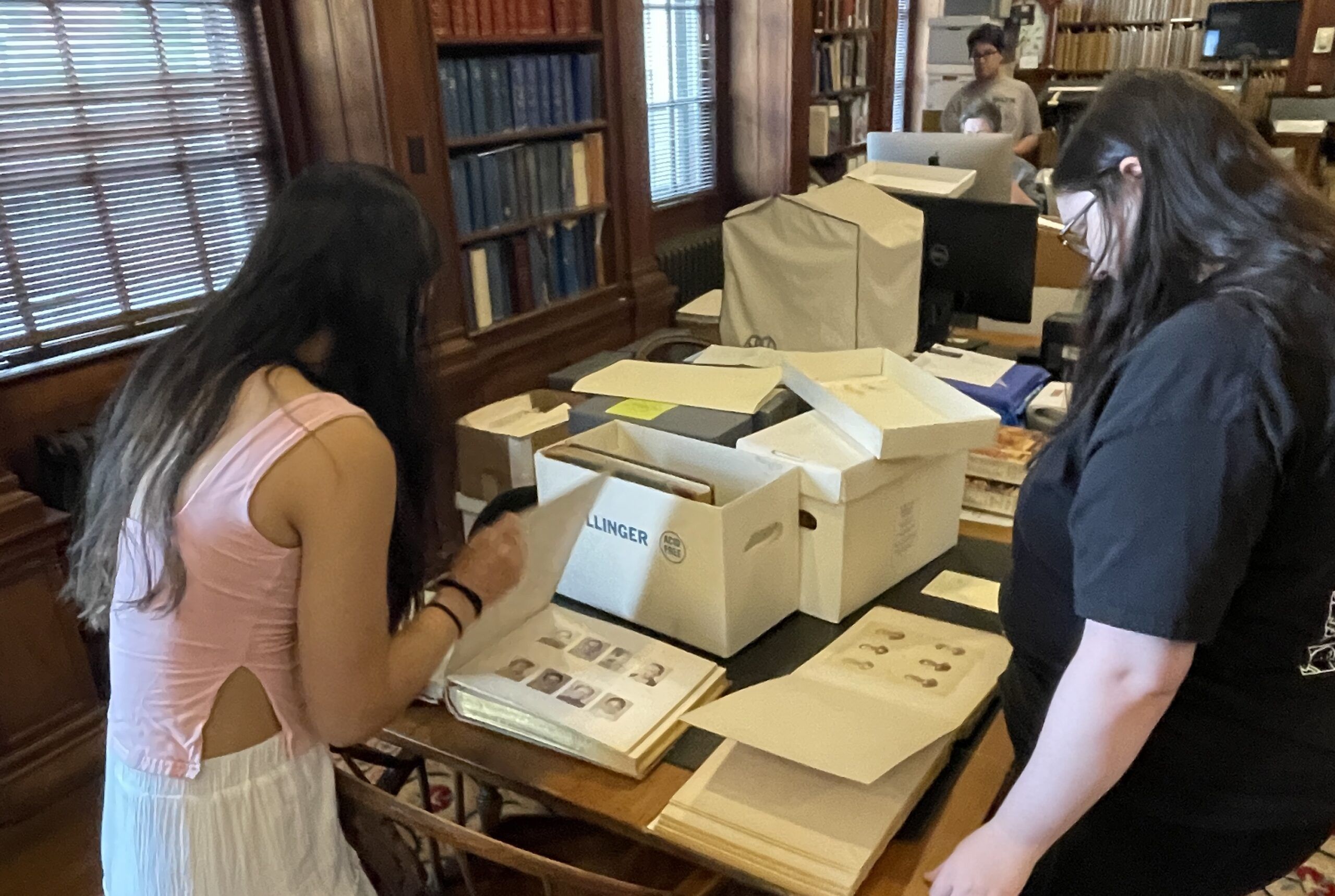 Two people examine archival photographs and documents on a table in a library or archive, possibly as part of the Summer Undergrad Research Grant Program, with boxes and bookshelves in the background.