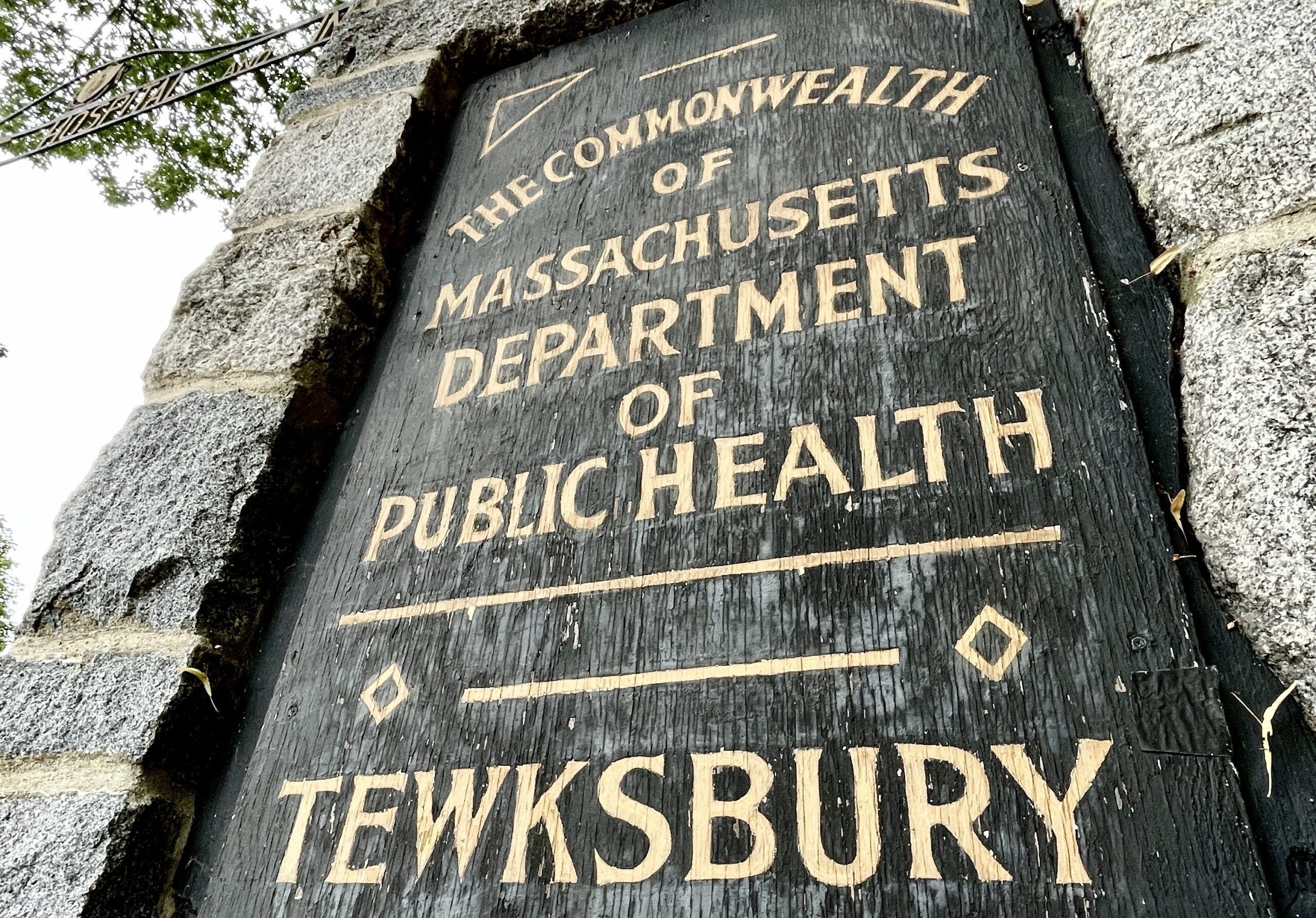 A weathered wooden sign reads "The Commonwealth of Massachusetts Department of Public Health, Tewksbury," mounted on a stone wall, home to the Summer Undergrad Research Grant Program.