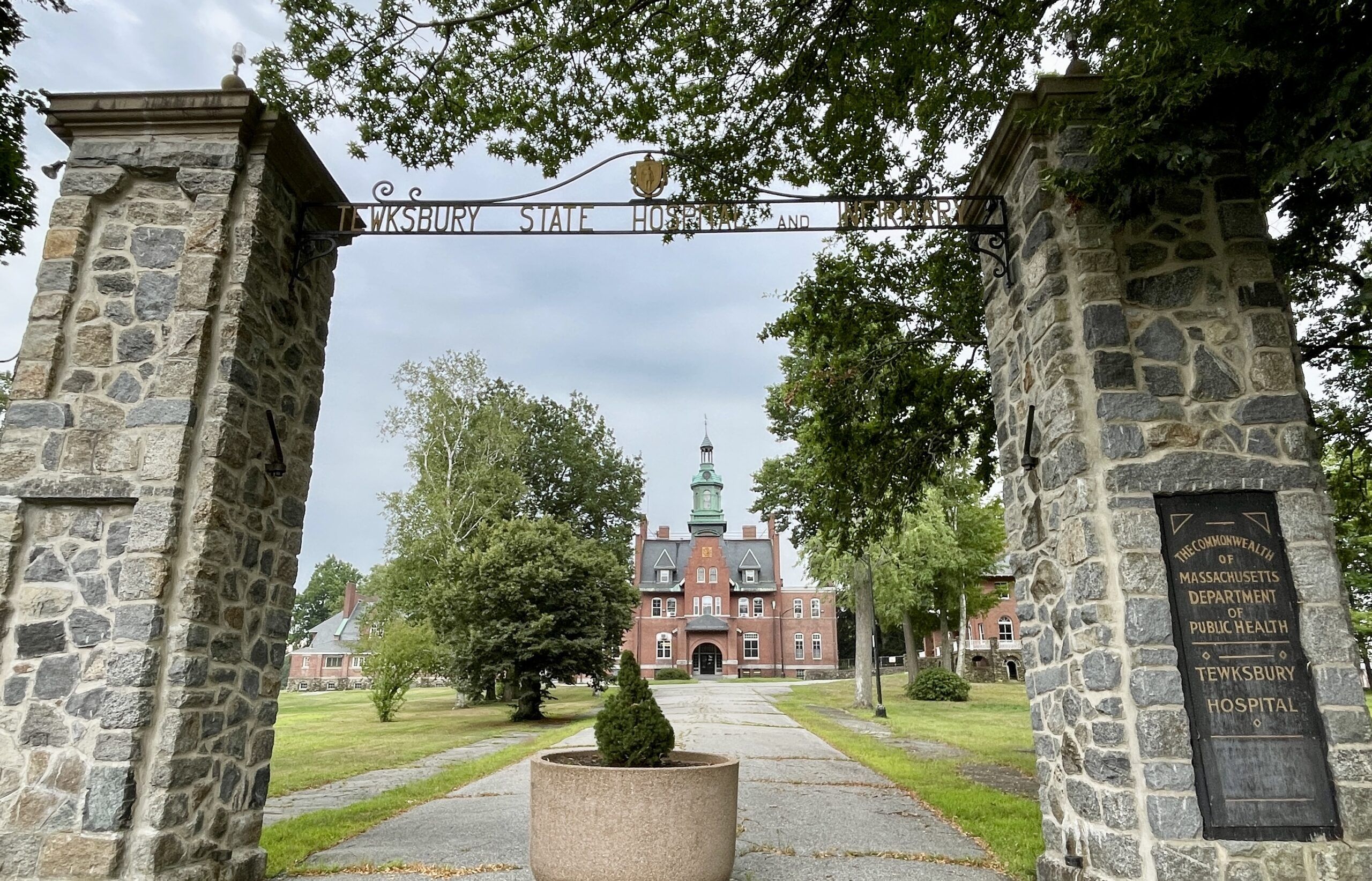 Stone entrance gate with a metal sign reading "Tewksbury State Hospital" leads to a red-brick building with a green-capped clock tower, home to the Summer Undergrad Research Grant Program, surrounded by trees and grassy lawns.