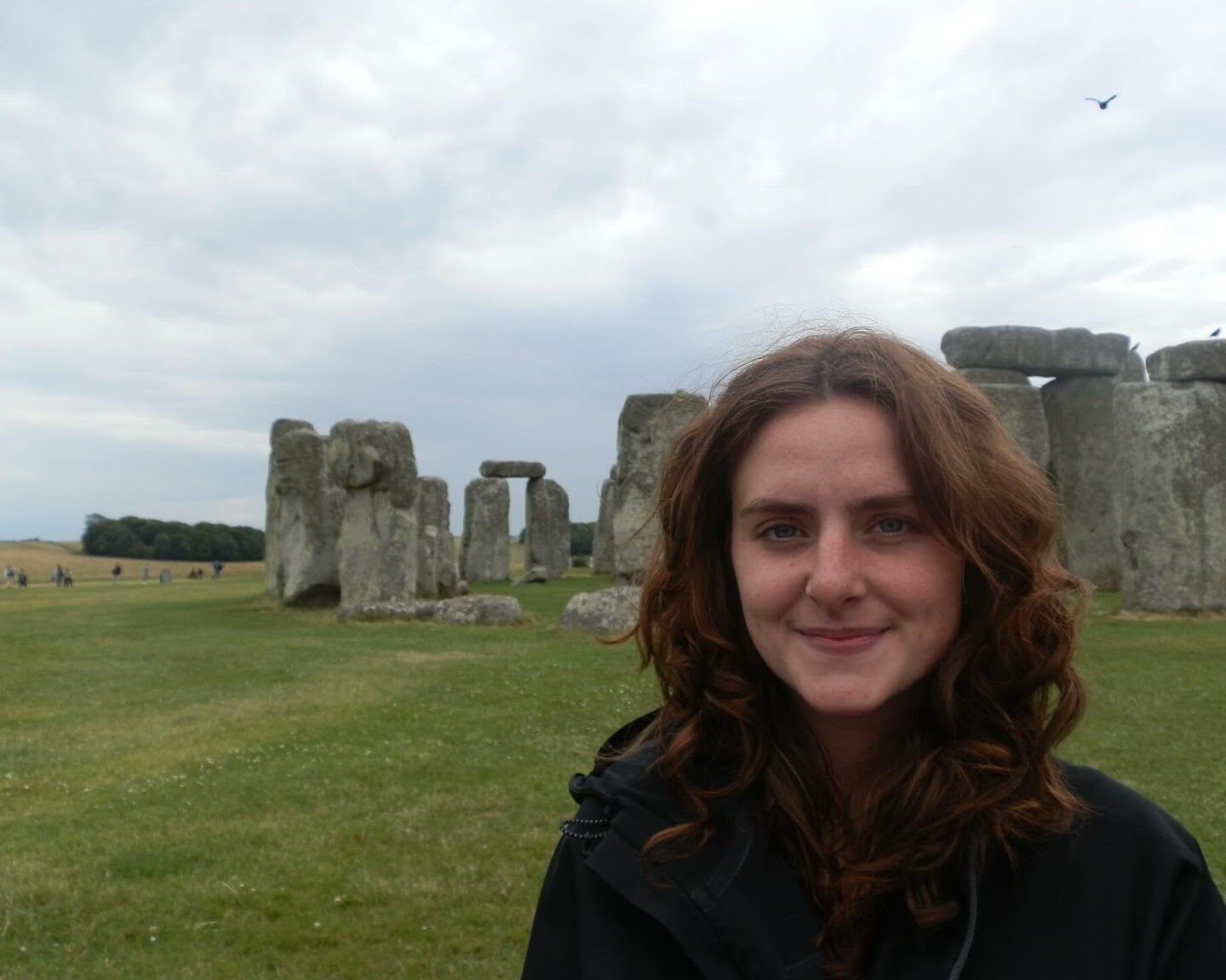 A person with long brown hair stands in front of Stonehenge on a cloudy day, with green grass and ancient stone structures visible in the background.