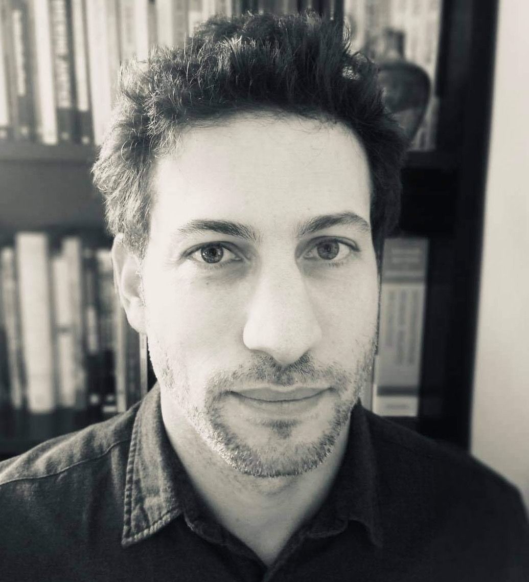 A man with short, curly hair and facial stubble looks directly at the camera. Bookshelves filled with books are visible in the background.