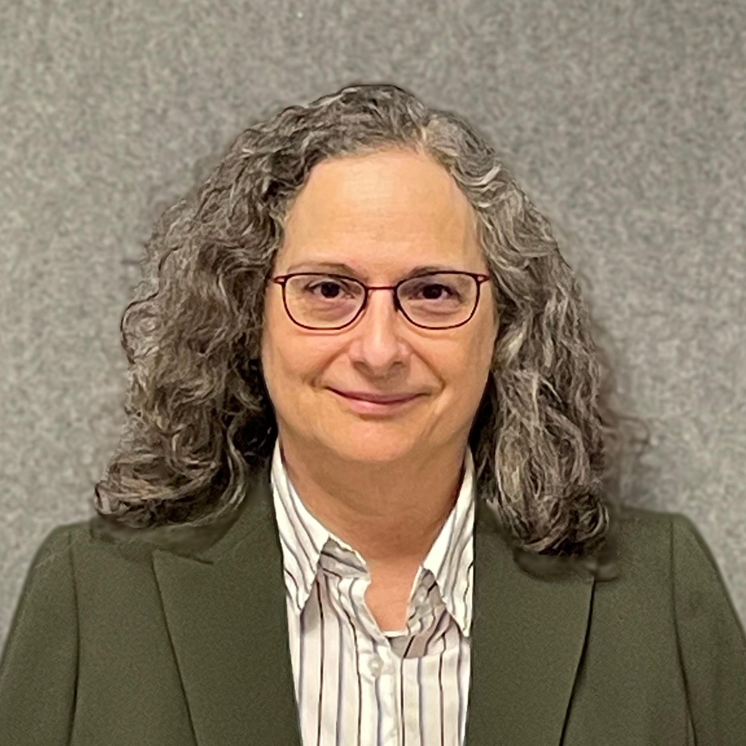 A person with curly gray hair, glasses, and a striped shirt under an olive green blazer stands in front of a gray textured background.