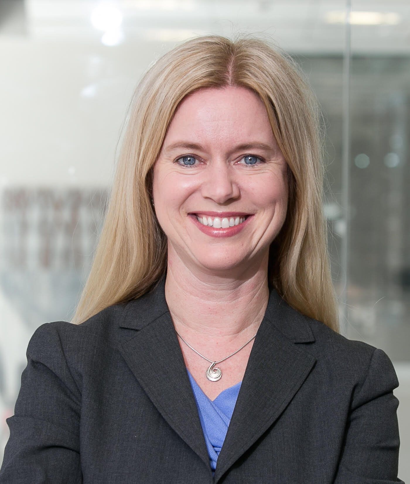 A woman with long blonde hair, wearing a dark blazer and blue top, stands smiling with her arms crossed.