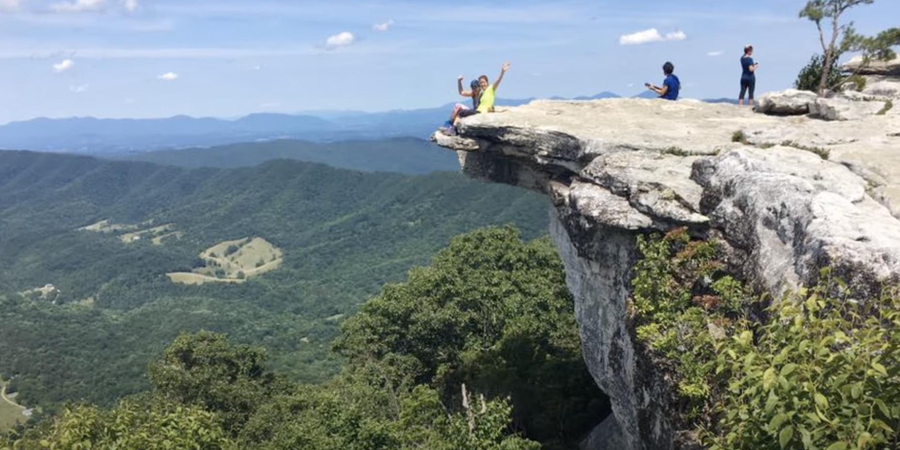 Two people wave from the tip of a mountain cliff surrounded by trees