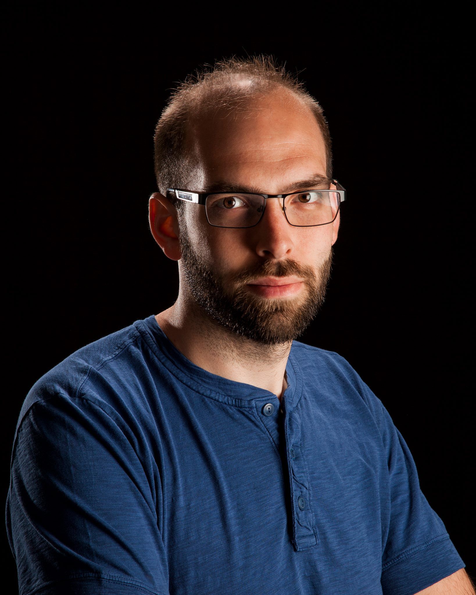 A man with glasses, a short beard, and short hair wearing a blue shirt looks directly at the camera against a black background, embodying the confident presence of Communication Faculty.