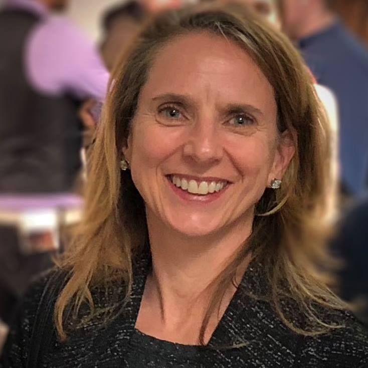 A woman with shoulder-length light brown hair, wearing a dark blazer and earrings, smiles at the camera. The background is blurred with people in a social setting, reflecting her role within the Communication Faculty.