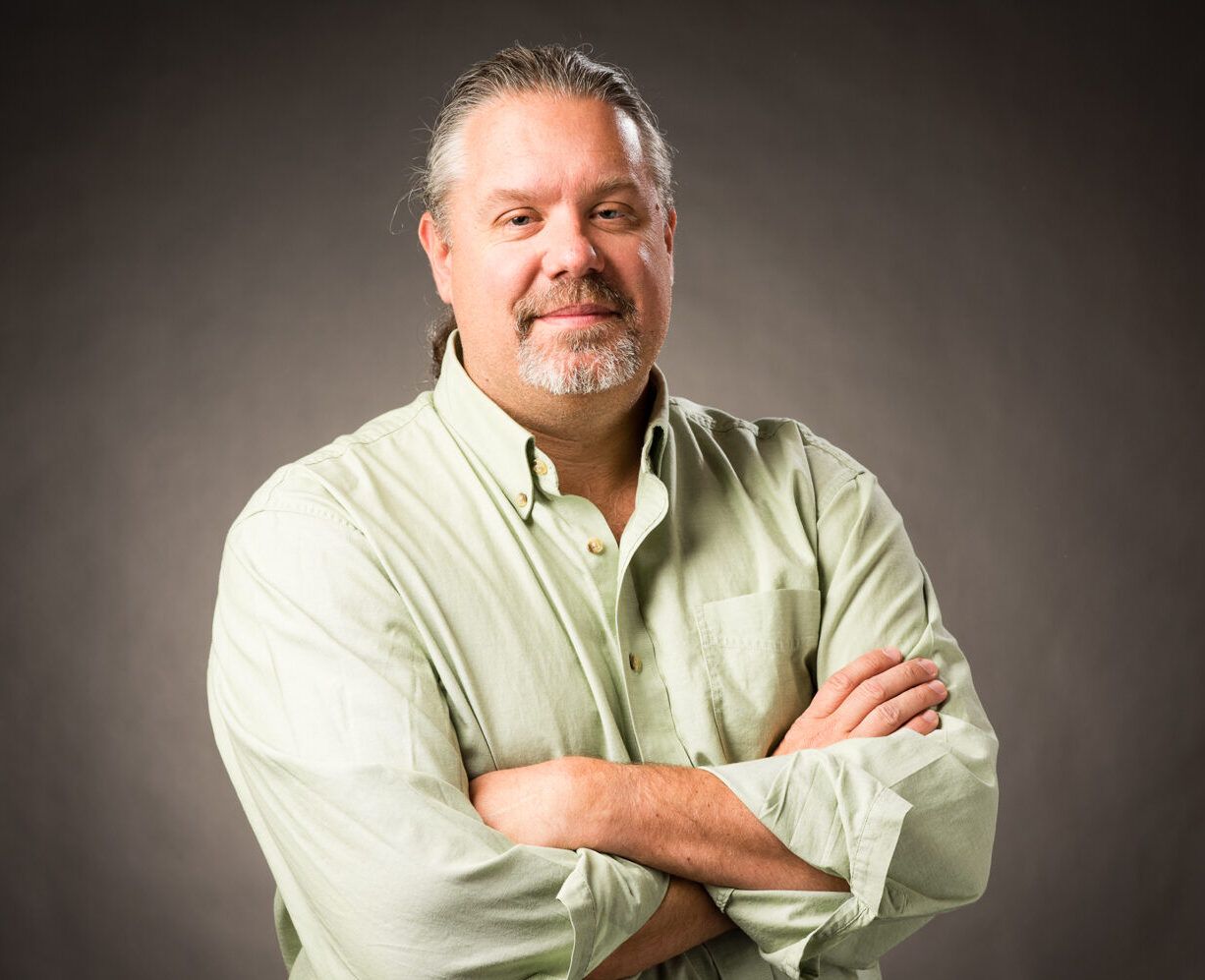 A middle-aged man with long hair and a beard, wearing a light green button-up shirt, stands with arms crossed against a plain dark background.