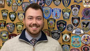 Portrait of Criminal Justice Worcester State alum Jared K. Richard standing in front of badge wall