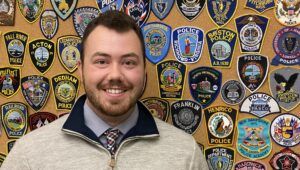 Portrait of Criminal Justice Worcester State alum Jared K. Richard standing in front of badge wall