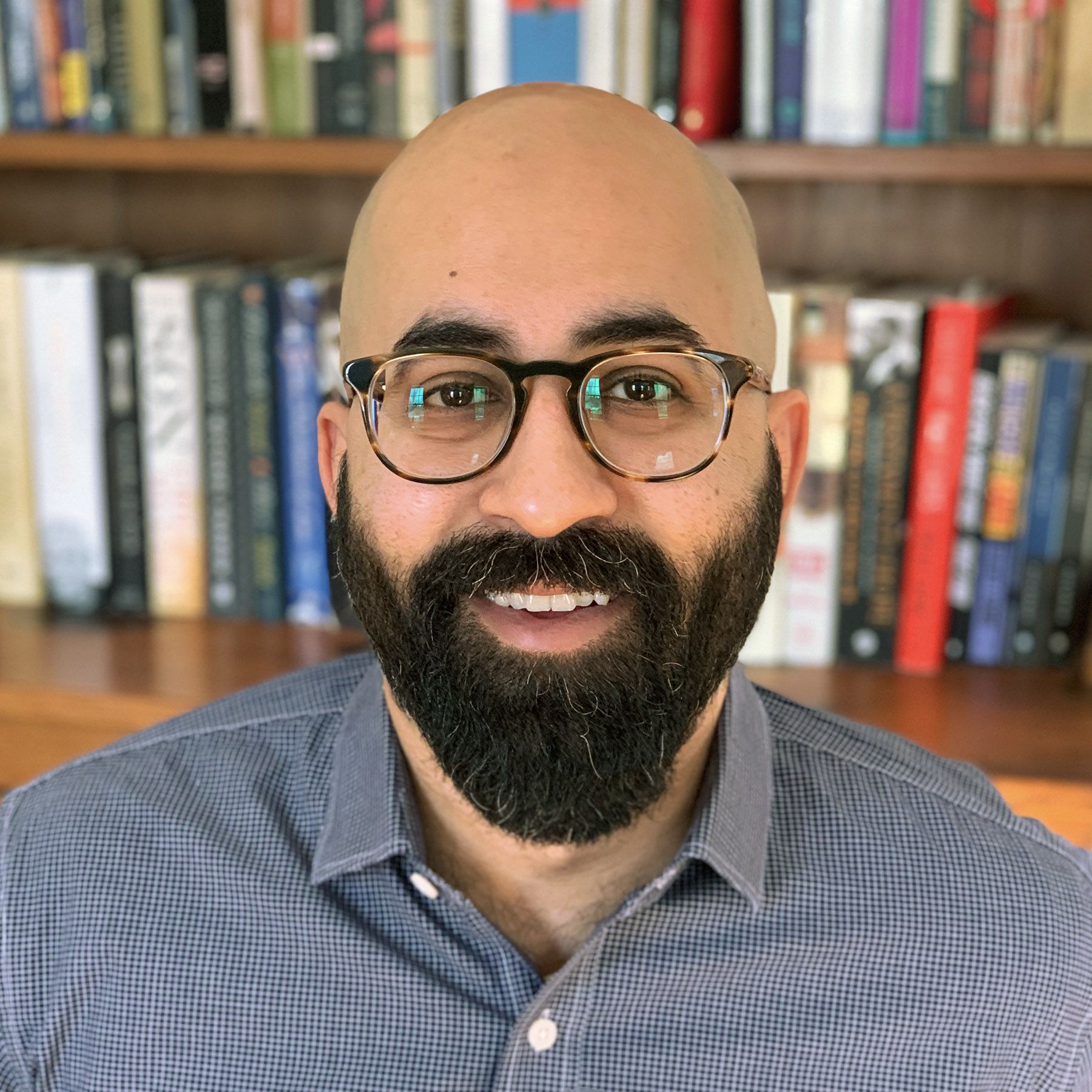 A man with glasses, a bald head, and a beard is smiling in front of a bookshelf filled with books.