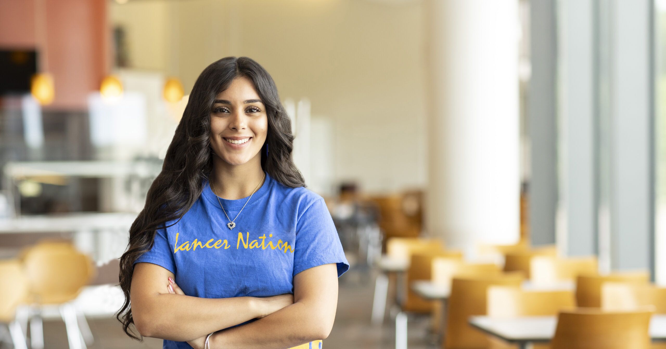 A woman with long dark hair, wearing a blue "Lancer Nation" t-shirt, stands with arms crossed in a bright, modern cafeteria with yellow chairs and large windows.