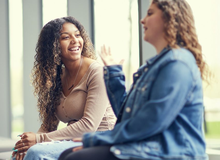 Two young women with curly hair sit indoors at Worcester State, engaged in conversation in español. One is smiling and listening, while the other gestures with her hand.