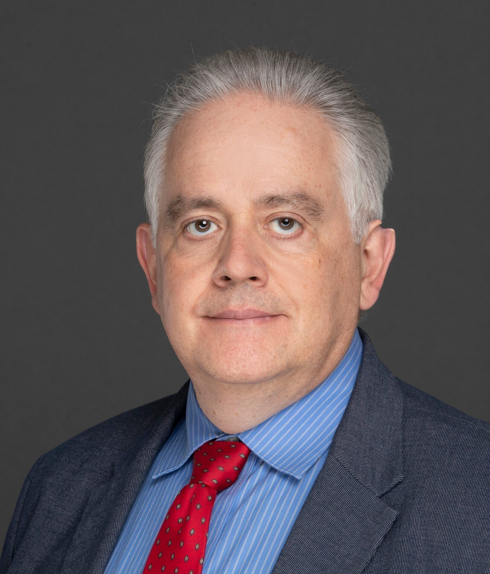 A middle-aged man with gray hair wearing a blue suit jacket, blue striped shirt, and red polka dot tie against a plain gray background.