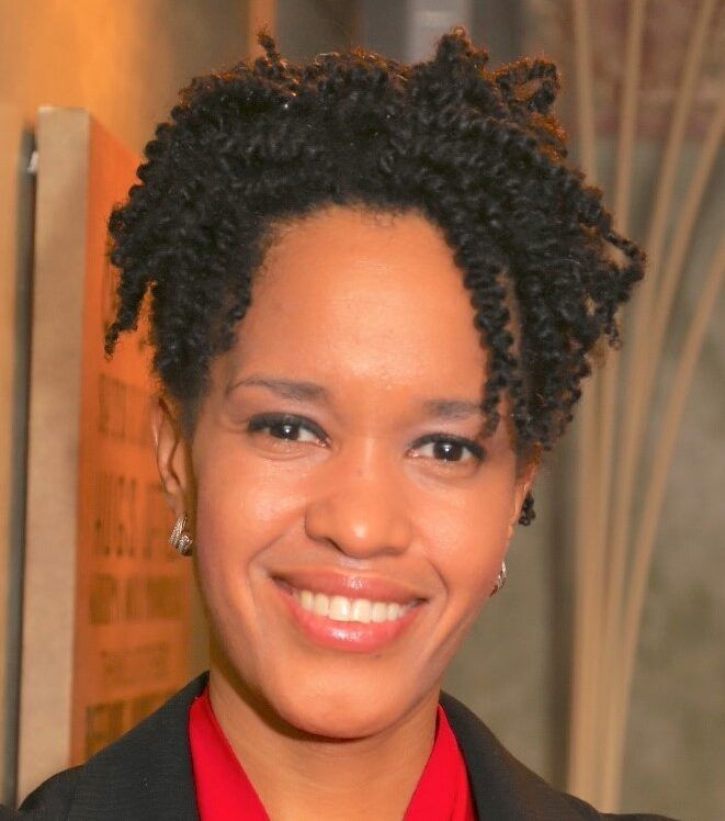 A woman with short curly hair, wearing a red blouse, black blazer, and necklace, smiles at the camera in an indoor setting, embodying the confidence and professionalism inspired by Title IX initiatives.