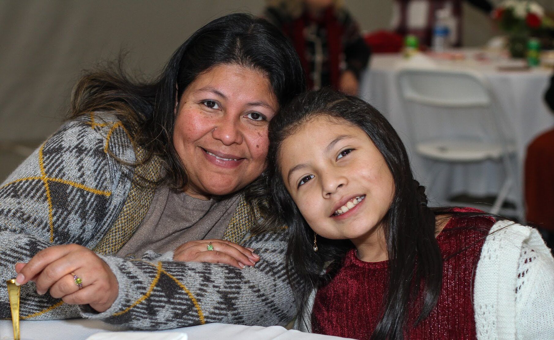A woman and a young girl sit closely together at a table, smiling at the camera. There are plates with food and a water bottle on the table.