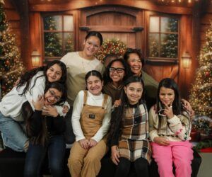 A group of eight people, including children and adults, pose and smile in front of a festive, holiday-themed backdrop with lights and wreaths.
