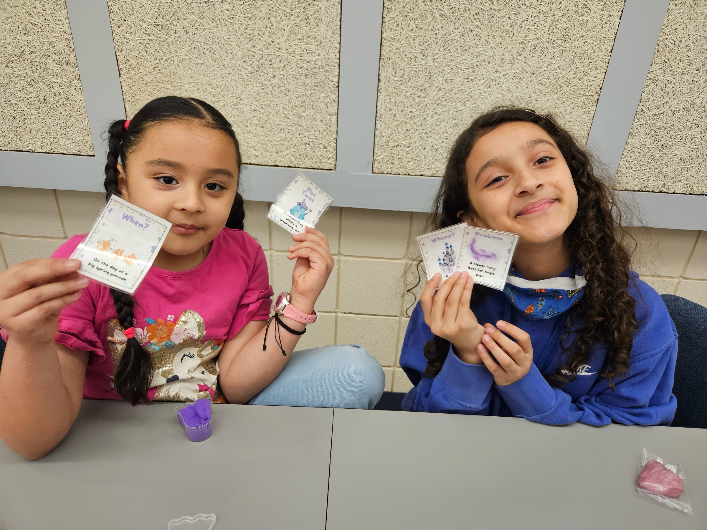 Two girls sitting at a table hold up hand-drawn cards and smile at the camera, with craft supplies on the table in front of them.