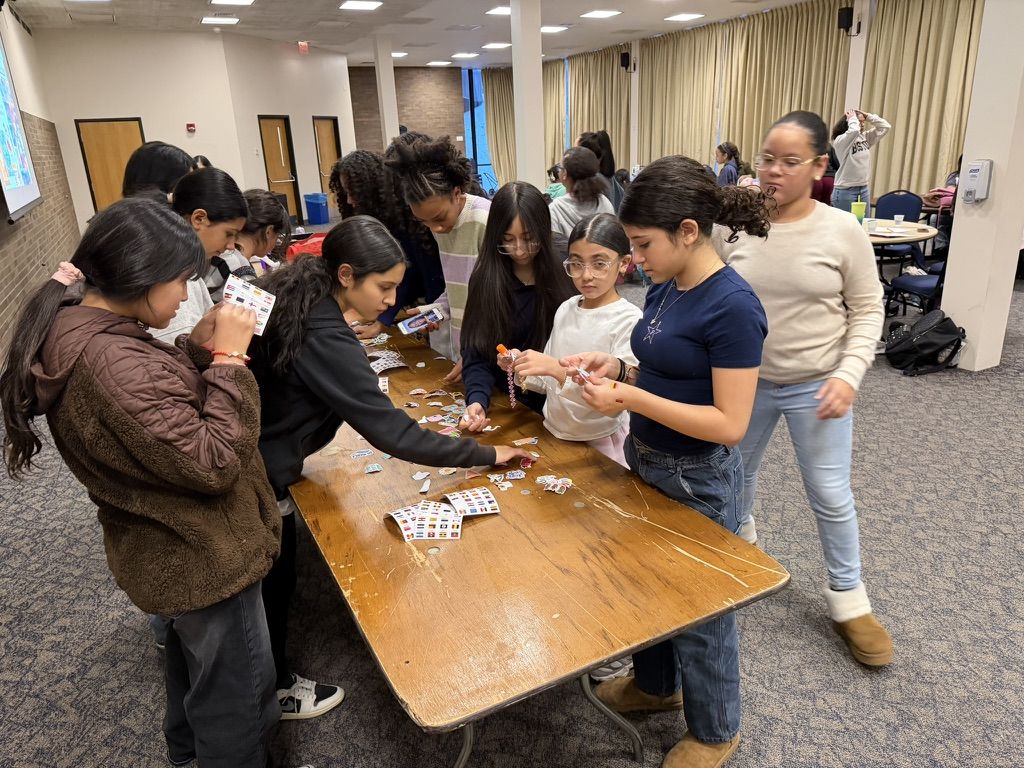 A group of girls gathers around a wooden table covered with stickers and small items, engaging in a craft activity in a classroom setting.