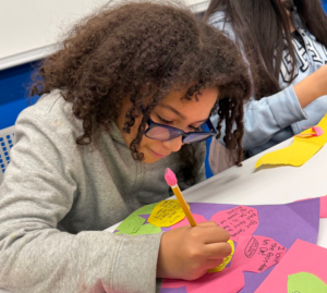 A child with glasses writes on a colorful paper in a classroom, surrounded by cut-out shapes and other students.