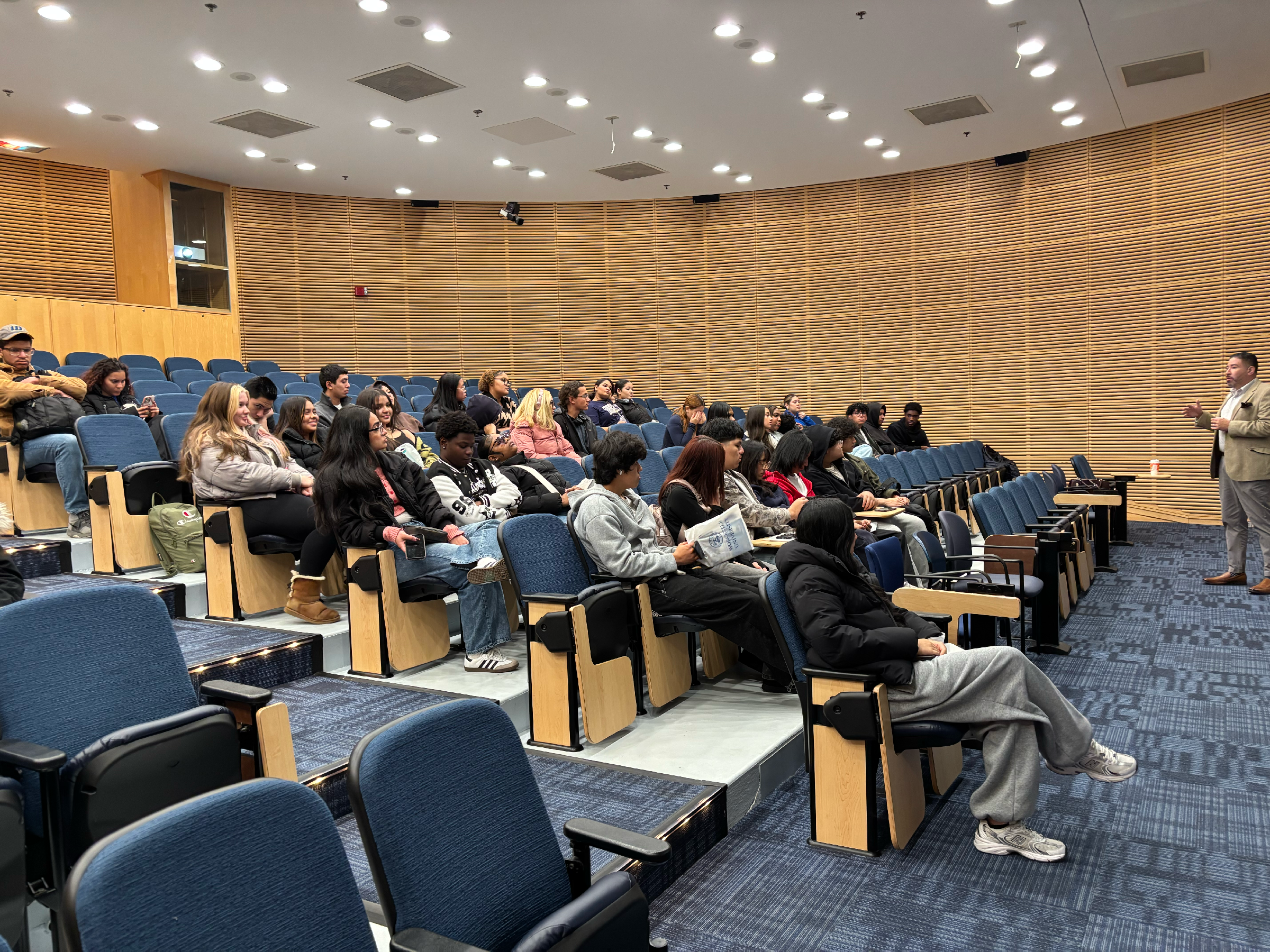 A group of people sit in an auditorium watching a standing speaker give a presentation. Some audience members hold papers or notebooks.
