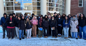A group of young adults stand together outside a building with large windows and columns on a snowy day, posing for a group photo.