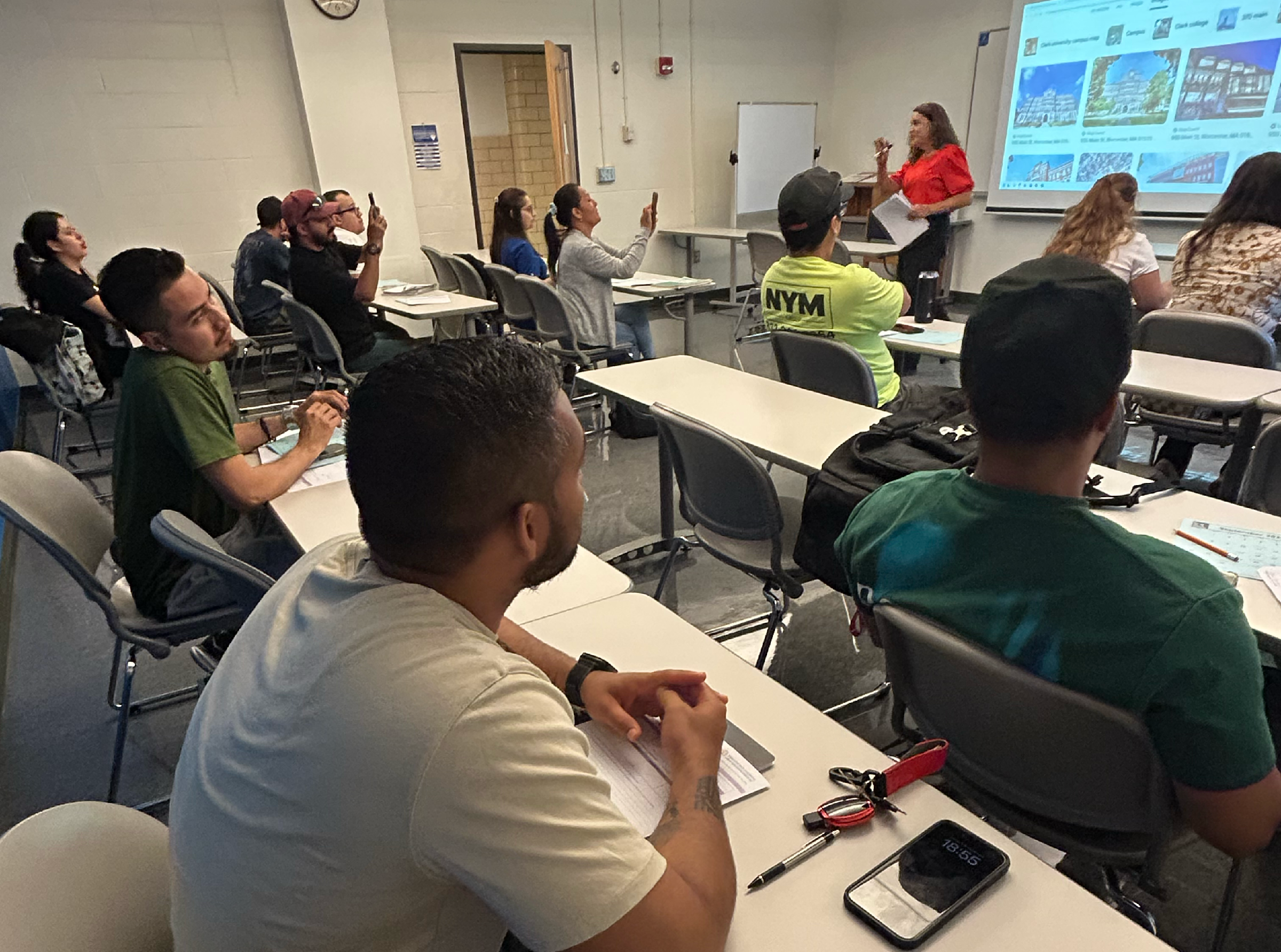 A teacher stands at the front of a classroom presenting slides to adult students sitting at desks while some take notes and others look at their phones.