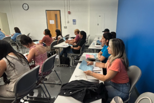 Adults sit at desks in a classroom, writing on papers and listening to someone outside the frame. A blue wall is visible to the right, and a clock hangs above a door at the back.