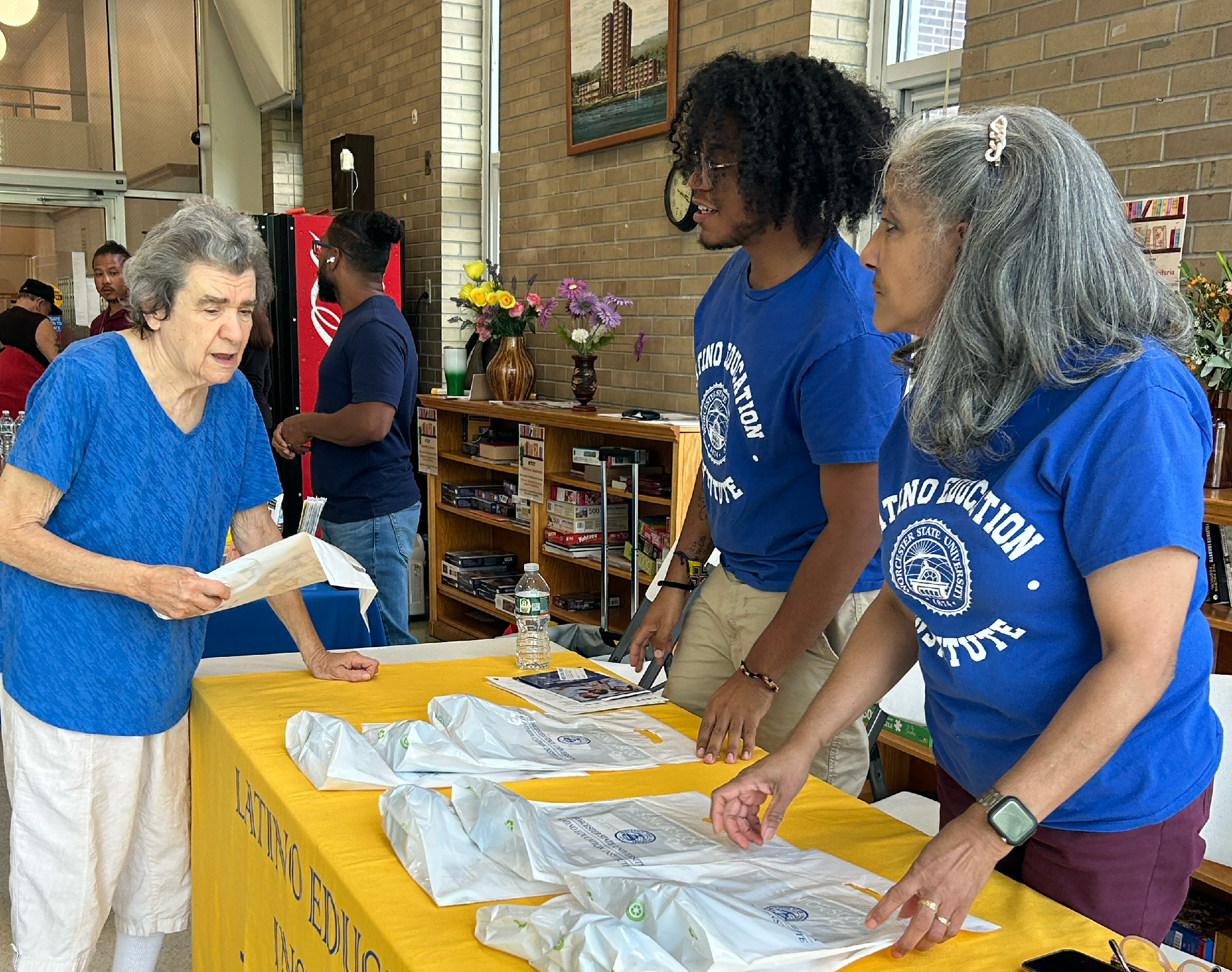 Two people in blue “Latino Education Institute” shirts assist an older woman at an information table with bags and papers inside a community center.