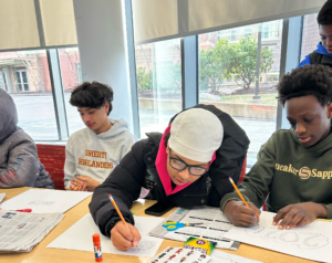 Three students sit at a table drawing on white paper with pencils, surrounded by school supplies, near a window with a view outside.