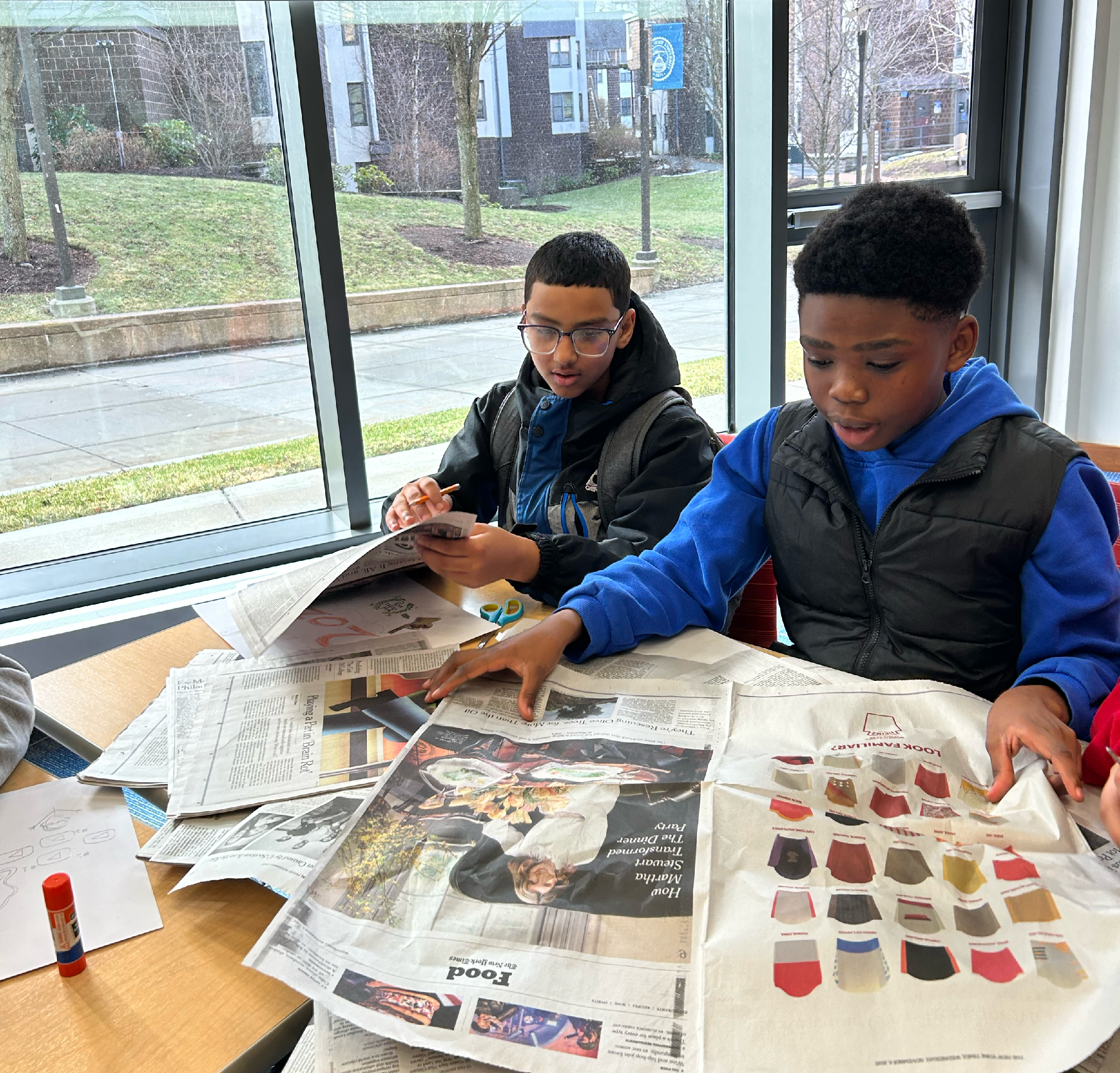 Two boys sit at a table covered with newspapers, cutting and arranging paper for a project near a large window on a rainy day.