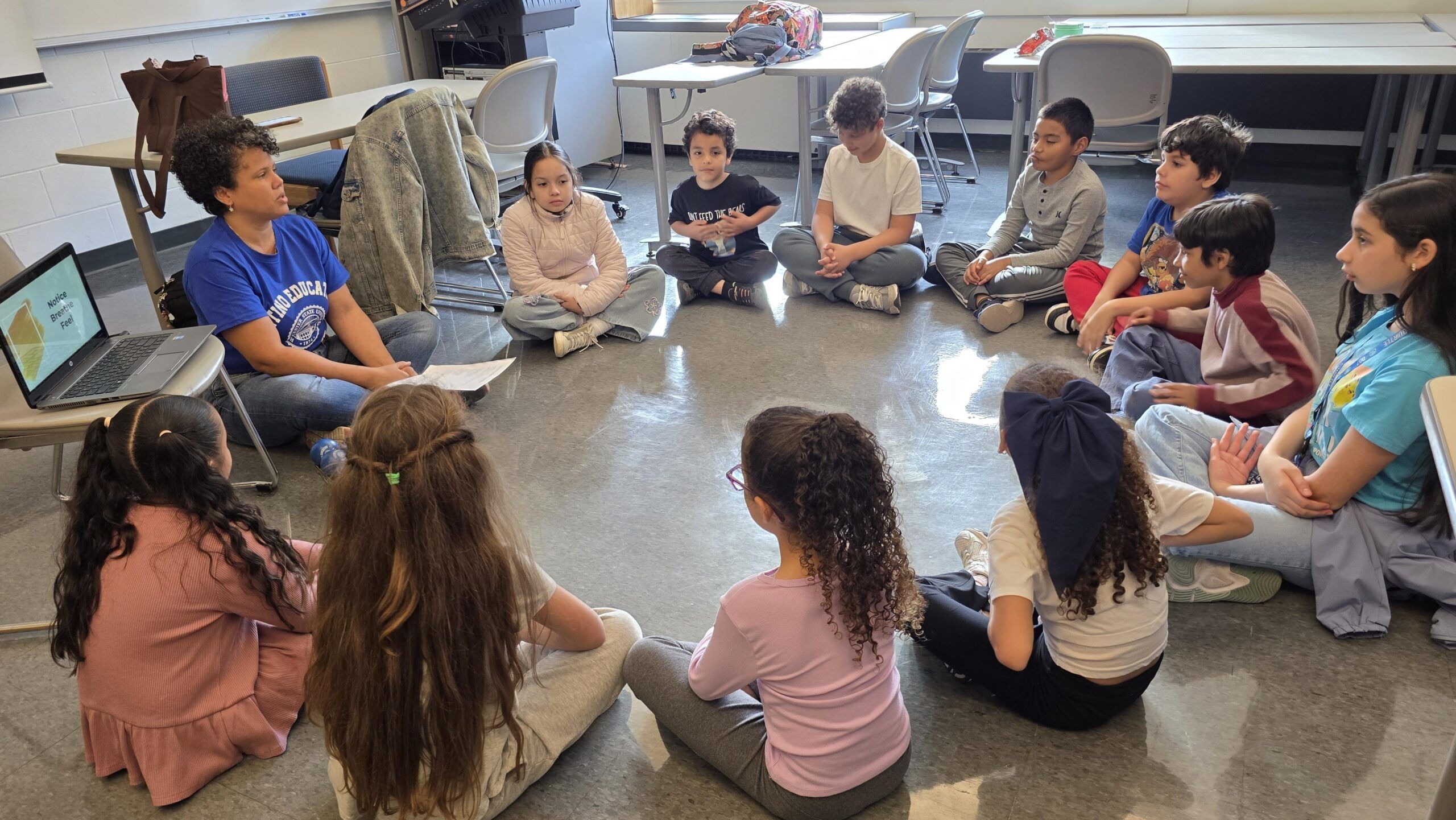 A group of children sit in a circle on the floor of a classroom, listening to an adult who holds a paper. A laptop is open nearby.