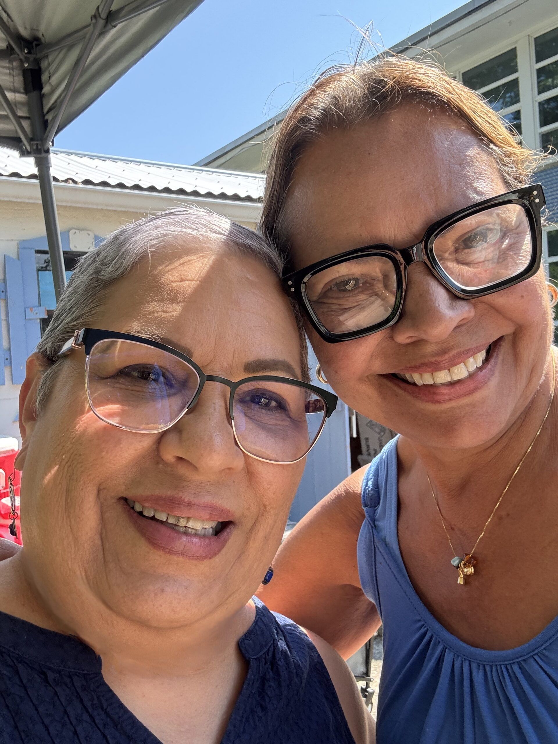 Two women with glasses smile at the camera outdoors on a sunny day, standing close together under a canopy. Their warm expressions reflect the camaraderie and inspiration often found through Latino Education Institute Leadership.