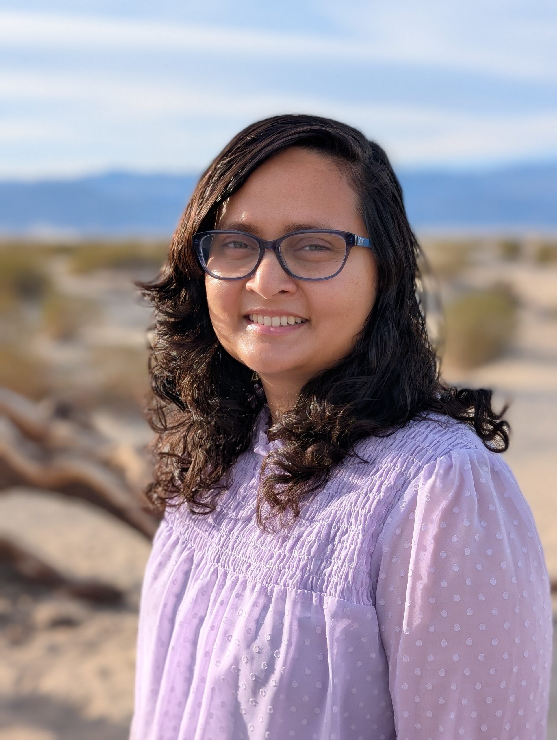 A person with glasses and curly hair smiles, wearing a light purple blouse. They stand outdoors in a desert landscape with dry shrubs and distant hills.