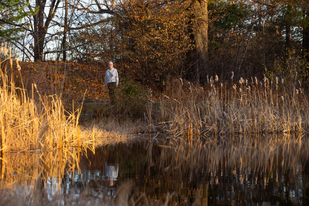 Judith Hoyer stands near a pond framed by tall grasses at Burncoat Park
