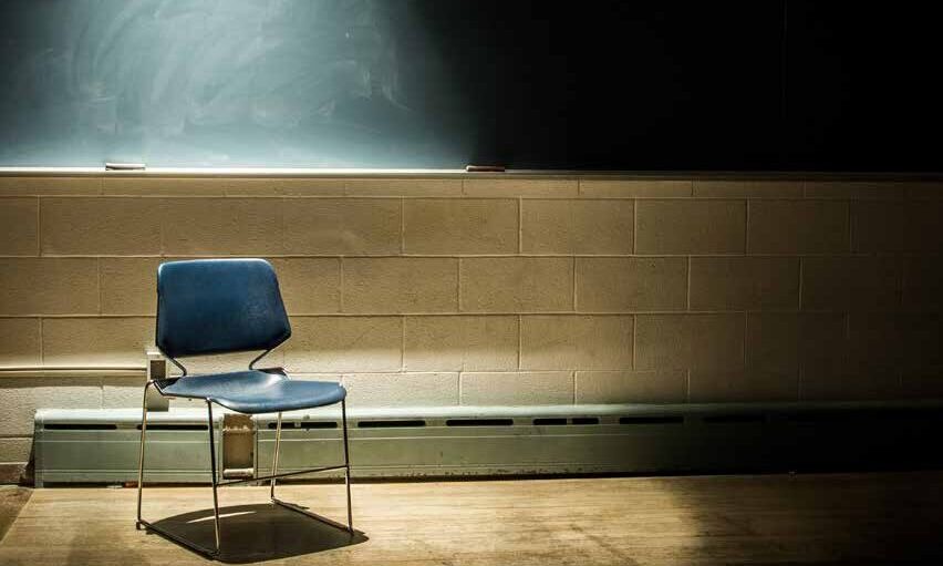 An empty classroom with a blank chalkboard and a metal chair under a spotlight