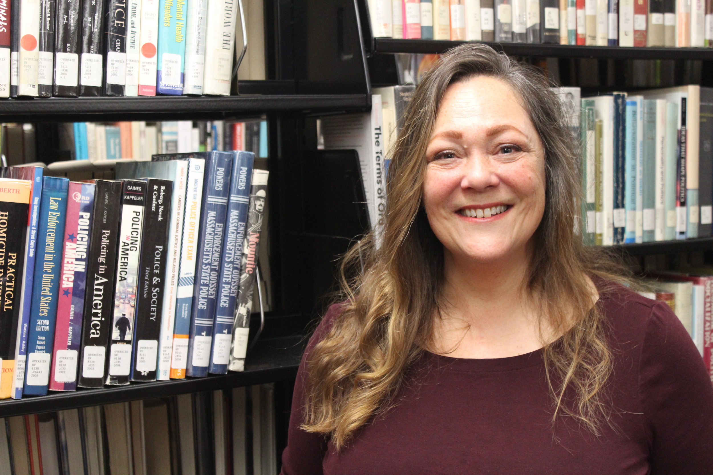 Professor Delaney stands next to the library stack where her book is shelved. She is looking at the camera and smiling.