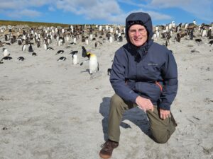 Hugh Donohue wears a windbreaker and kneels in beach sand. A colony of penguins is behind him.