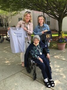 Flo Scarpaci sits in a wheelchair and wears a sharp navy suit. Behind her stand Sarah Bjorn and Jenna Beahn, two young, blond women. Sarah holds up a t-shirt saying Worcester State Lancers Alumni.