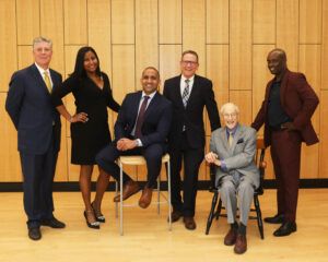 Six people, five standing and one seated, pose together in formal attire against a wood-paneled wall.