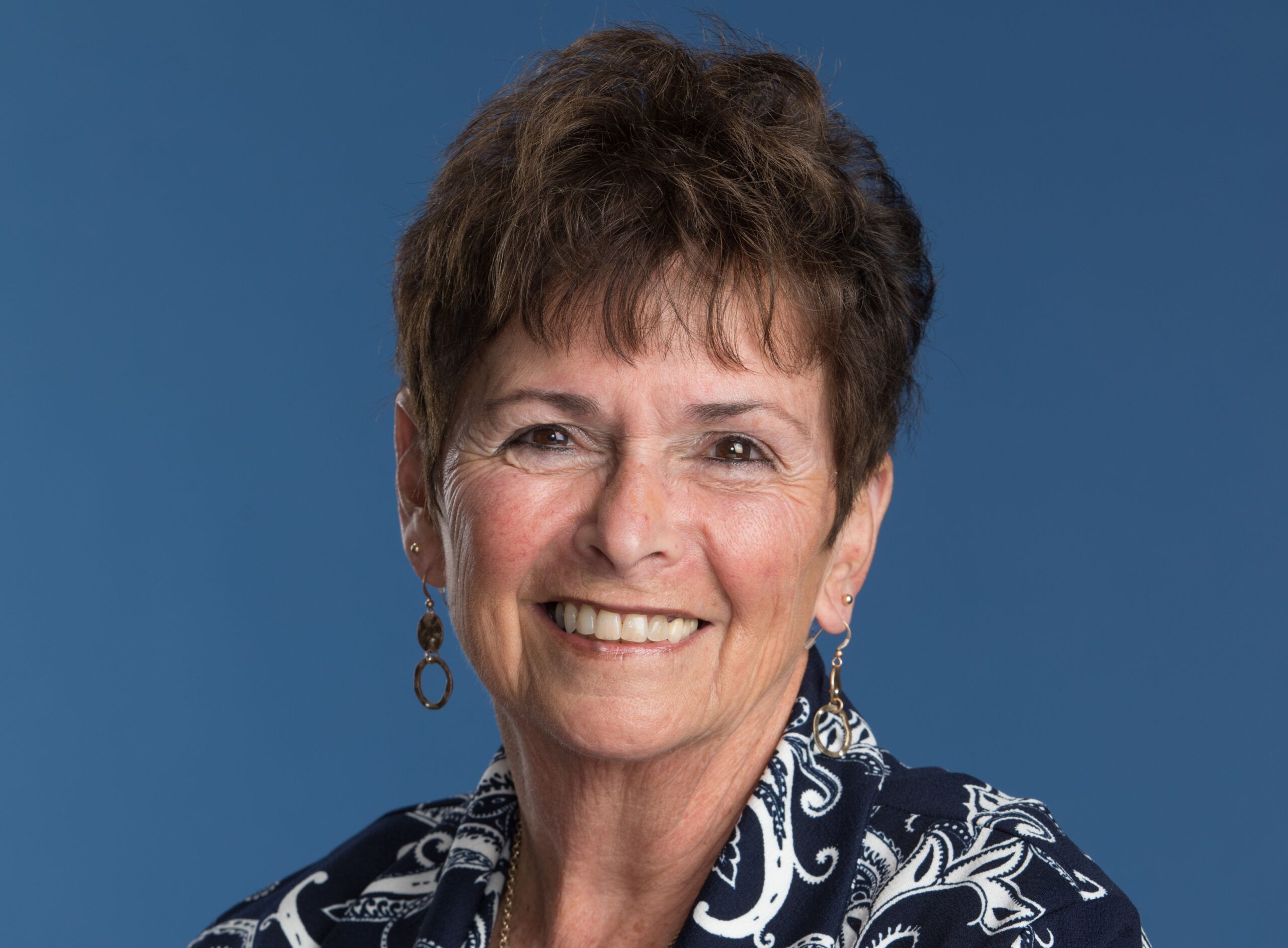 Smiling older woman with short brown hair, wearing hoop earrings and a navy blue patterned top, posed against a solid blue background.