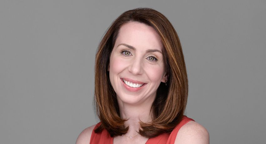 A woman with straight, shoulder-length brown hair, wearing a sleeveless coral top, smiles at the camera against a plain grey background.