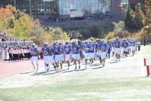 A college football team in blue and white uniforms runs onto the field, with spectators watching from the stands in the background.